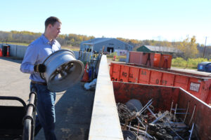 Man holding metal tire rim approaching large metal recycling bin.