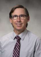 A headshot of a Caucasian man with straight, dark brown hair and brown eyes.