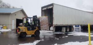 A forklift operator lifts a pallet of cardboard cases wrapped in plastic film into an open semi truck.