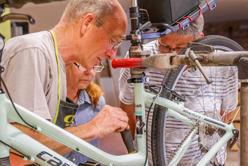 A person using a hand tool on a bicycle while two other people watch his work closely